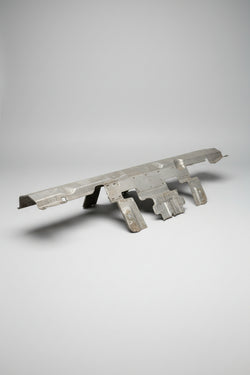 A used, unpainted metal automotive body panel or trim piece with mounting holes, resting on a blue surface inside a cluttered garage or workshop.